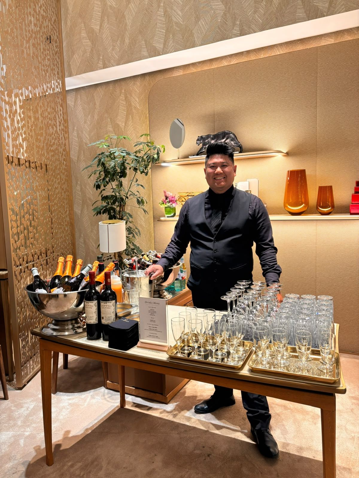 Barkeeper in schwarzer Weste hinter einem Tisch mit Sektgläsern auf goldenen Tabletts, Eiskübeln und Getränkekarte