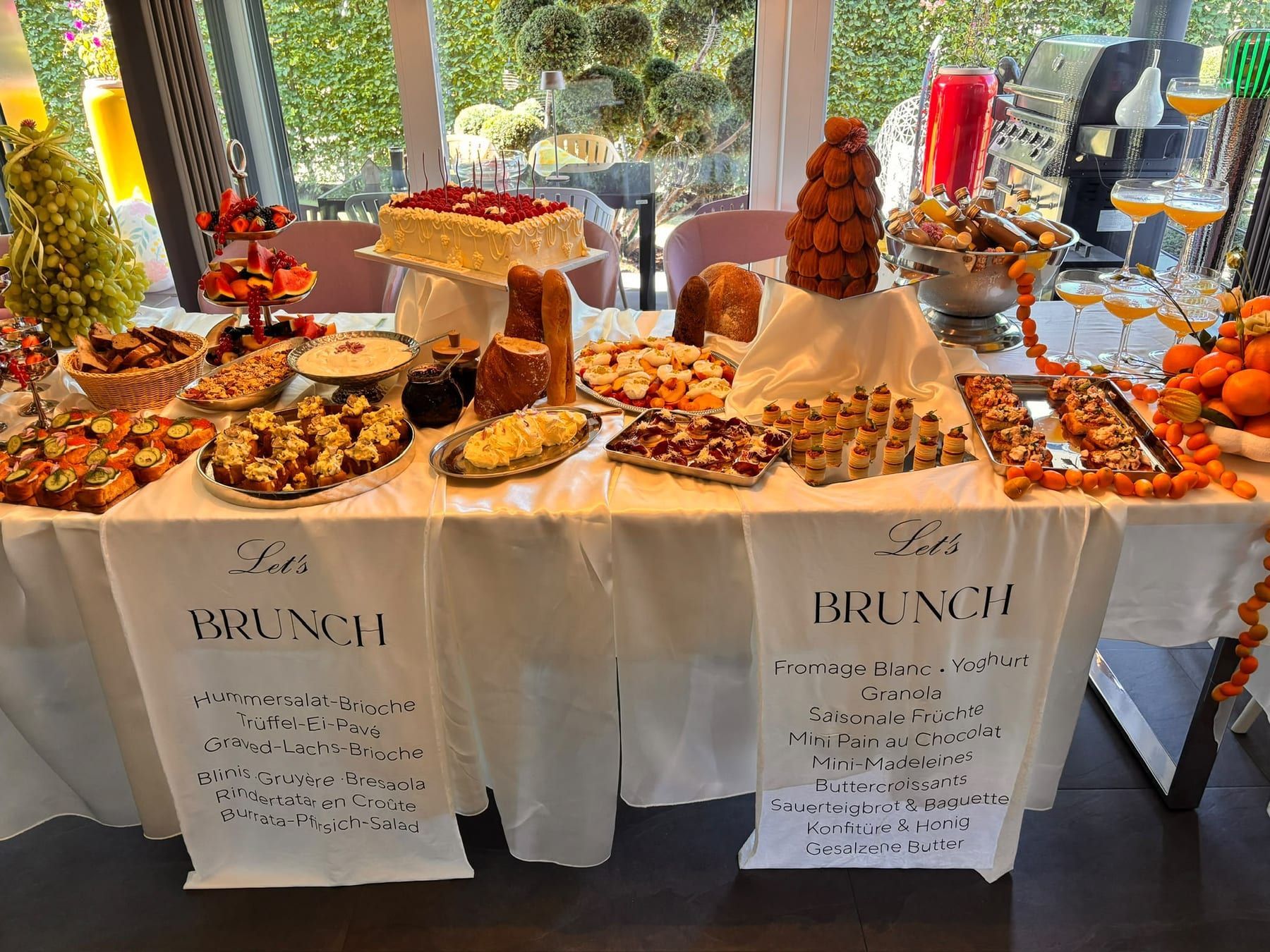 Brunch-Buffet mit Kuchen, Obstetagere, Brioche-Häppchen, Madeleine-Turm und Orangen auf einer weißen Tafel