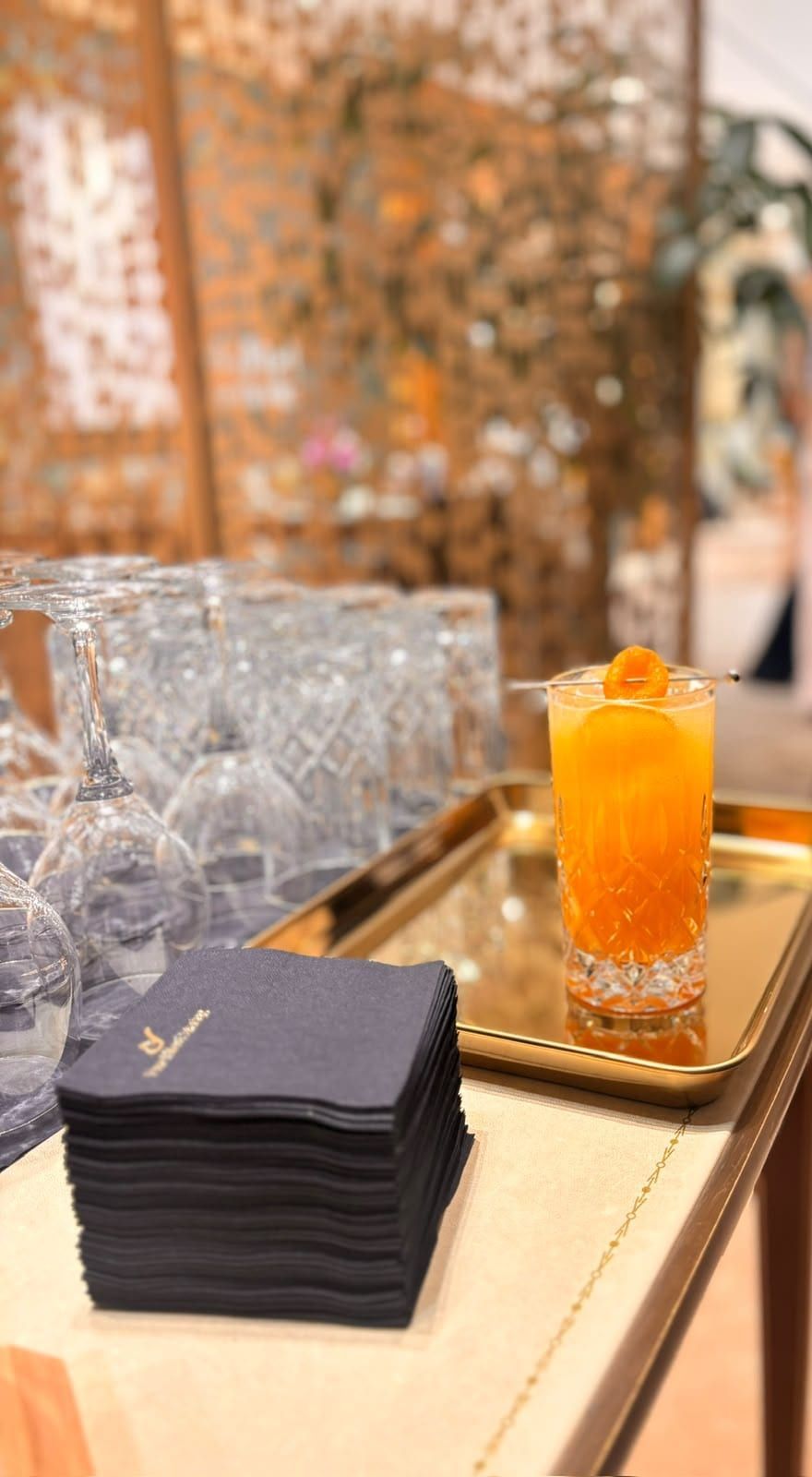 Orangefarbener Cocktail im Kristallglas auf einem Goldtablett, daneben ein Stapel dunkler Servietten