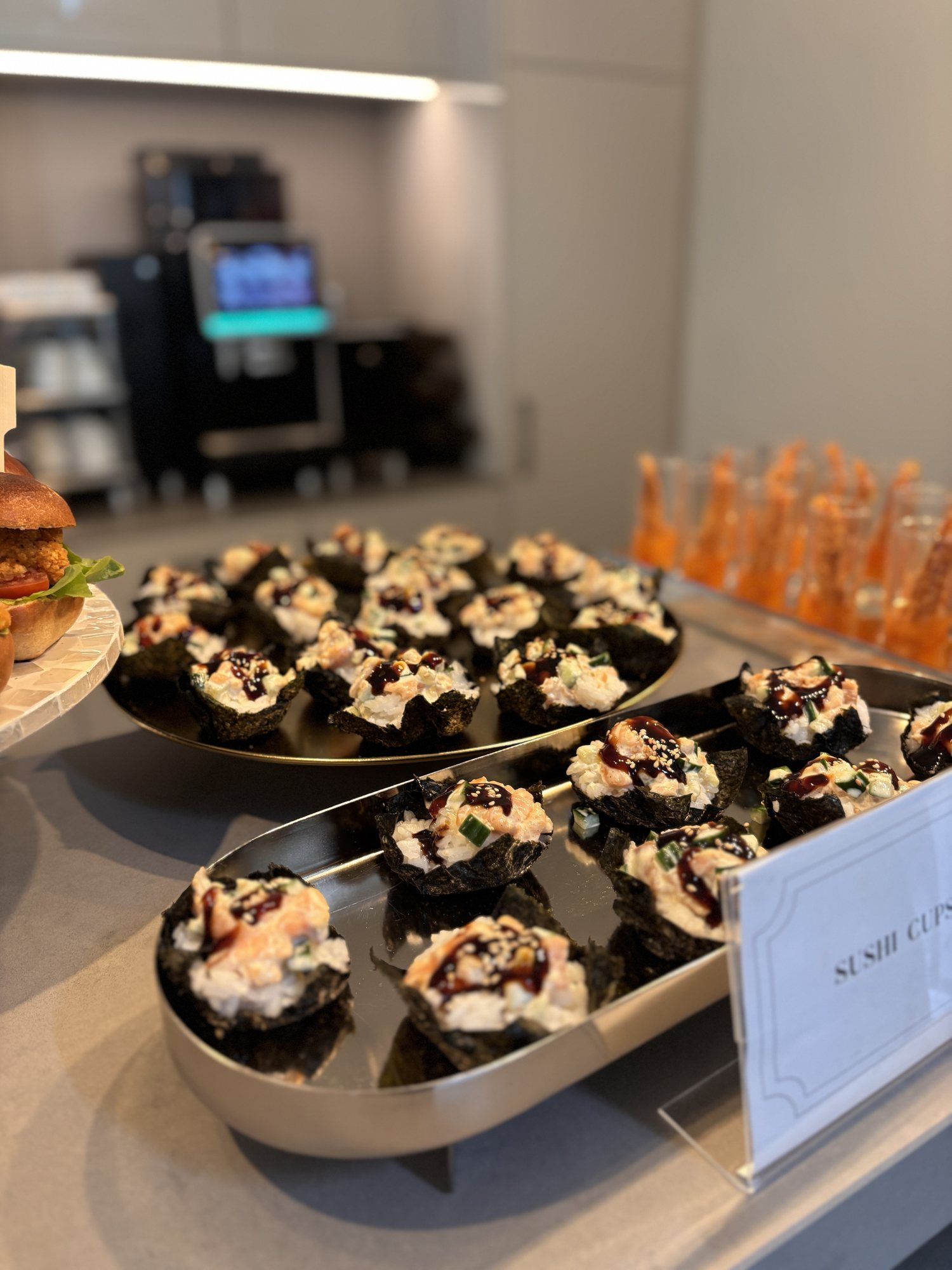 Sushi-Cups in Noriblättern auf Metallplatten, dahinter eine Kaffeemaschine