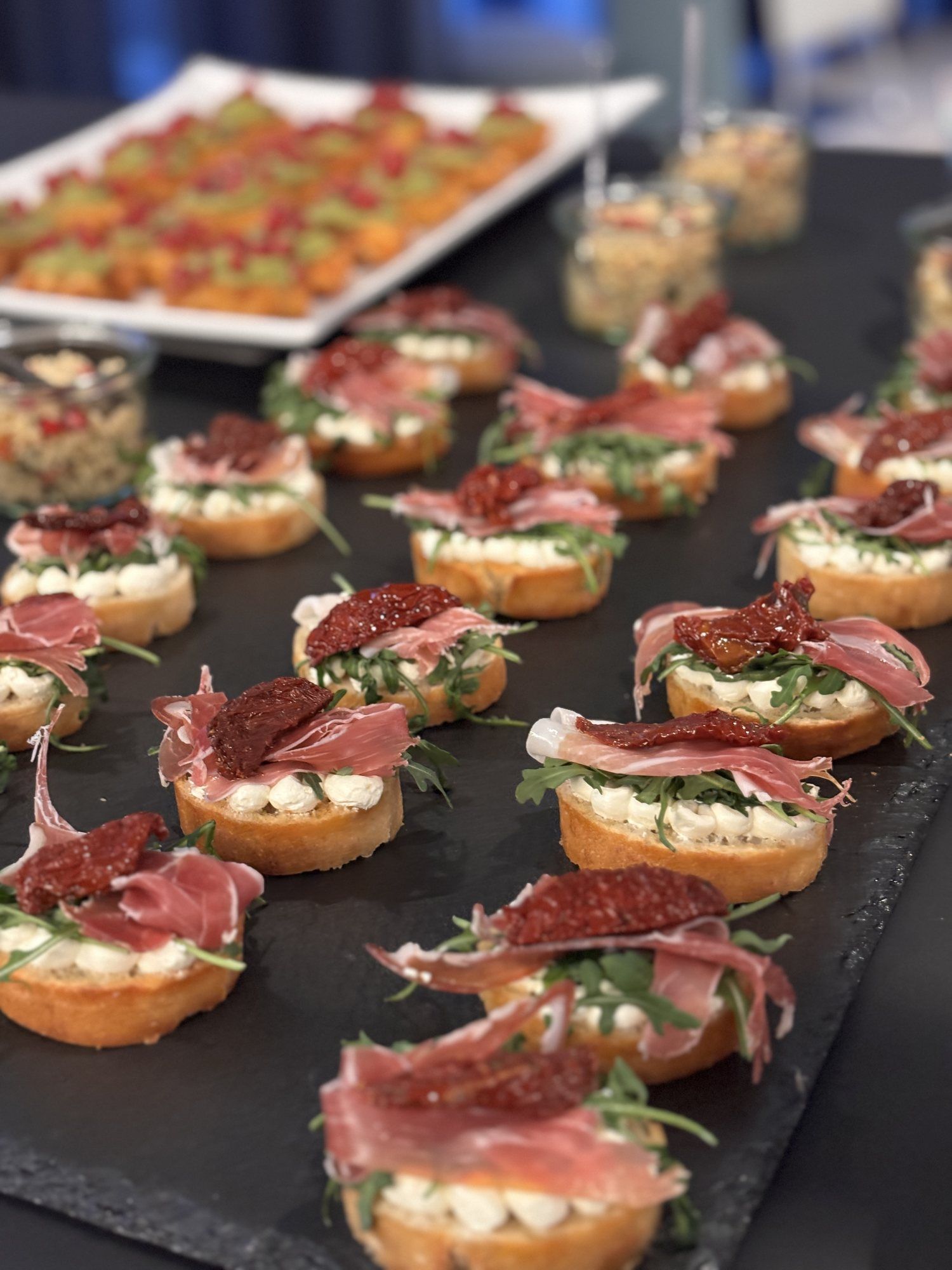 Röstbrotscheiben mit Frischkäse, Rucola, Schinken und getrockneten Tomaten auf einer Schieferplatte