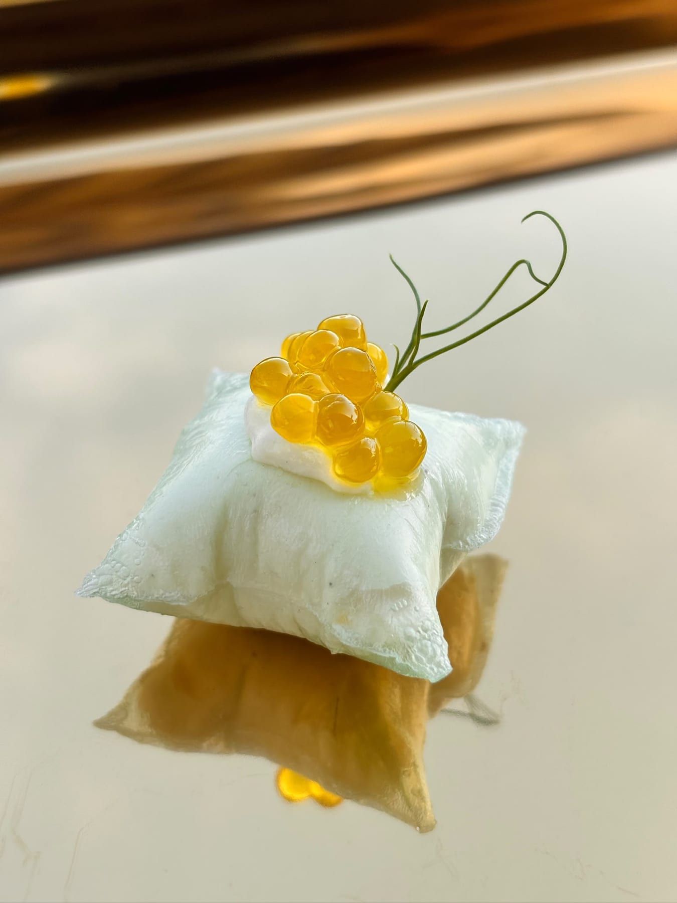 Kissenförmiges Häppchen mit gelben Perlen und Kräuterhalm auf einer Spiegelplatte